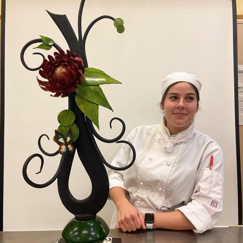 Woman in chef whites beside an ornate chocolate sculpture